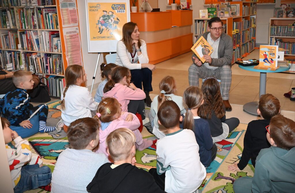Bibliotekarka Izabela Szczyż i Krzysztof Polak siedzą przed dziećmi. Krzysztof Polak trzyma w ręku książkę 