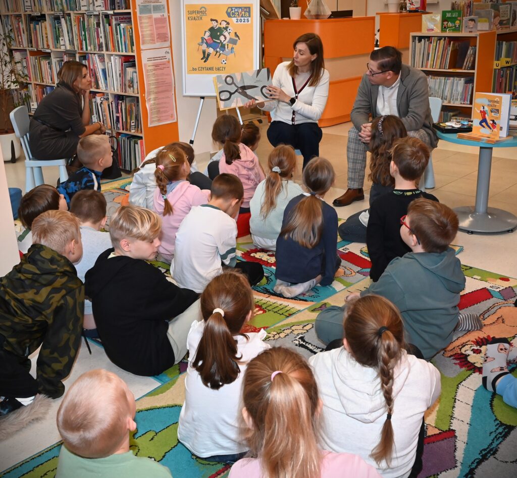 Bibliotekarka Izabela Szczyż pokazuje dzieciom książkę. Obok siedzi Krzysztof Polak