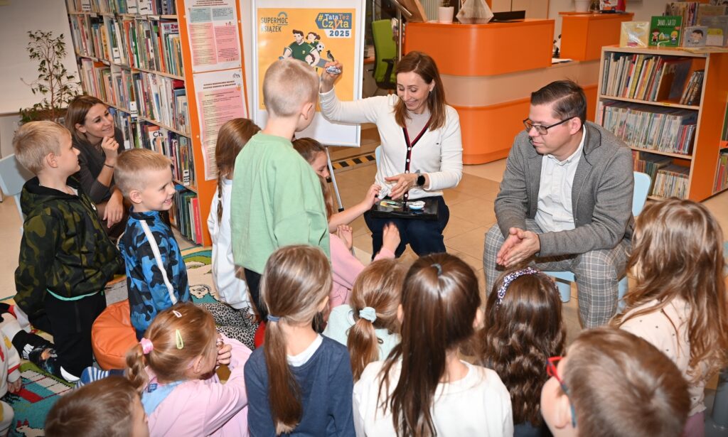 Bibliotekarka Izabela Szczyż pokazuje pomalowany kamień dzieciom. Obok siedzi Krzysztof Polak