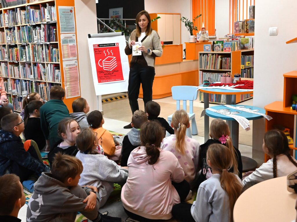 Bibliotekarka Izabela Szczyz stoi przed siedzącymi na dywanie dziećmi z książką w ręku.