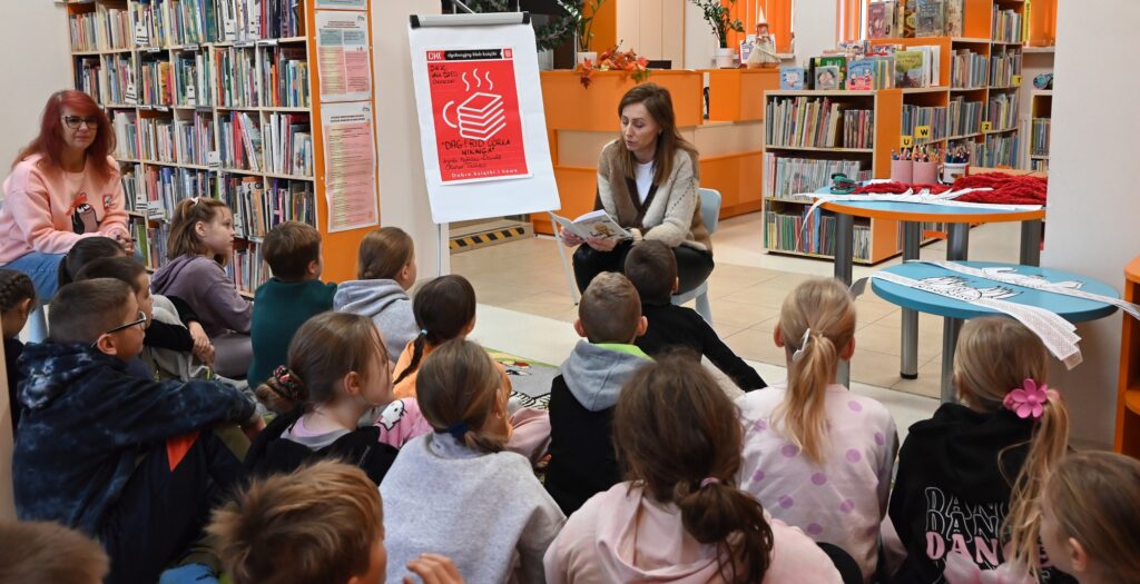 Bibliotekarka Izabela Szczyz siedzi na małym krzesełku przed siedzącymi na dywanie dziećmi i czyta im książkę.