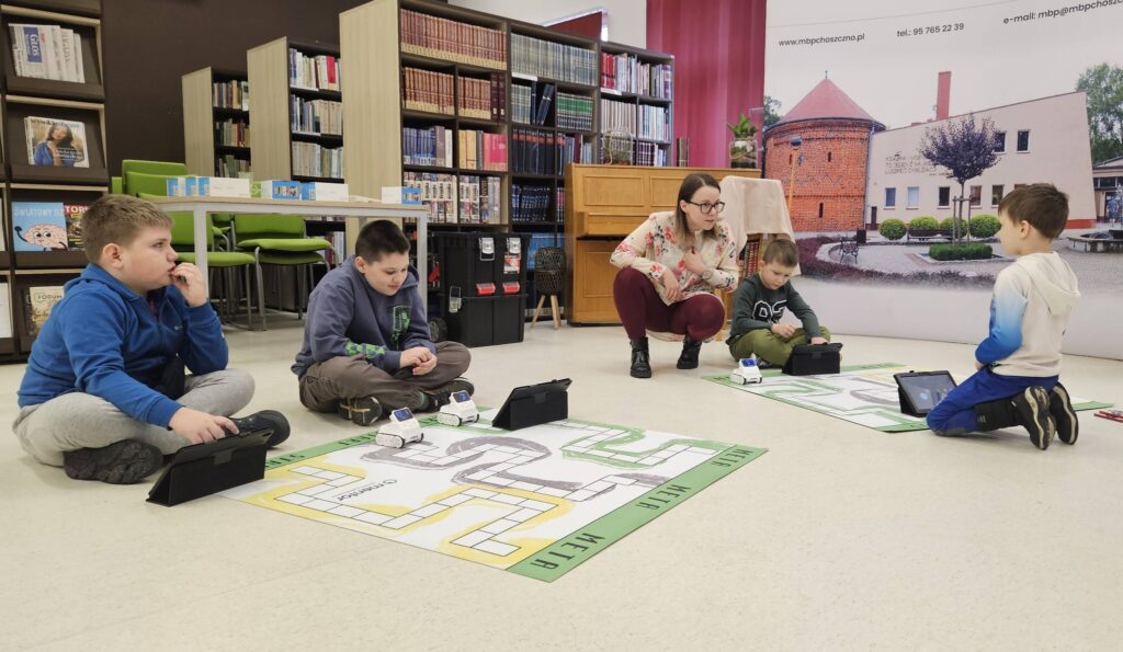 Na podłodze leżą maty z narysowaną drogą. Przy nich siedzą po dwóch chłopcy. Mają tablety i roboty. Kobieta kuca i wyjaśnia jednemu z nich .