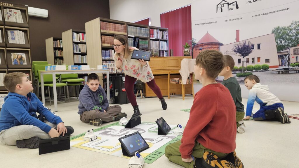 Na podłodze leżą maty z narysowaną drogą. Kobieta stoi pochylona z tabletem w rękach, coś tłumacząc, obok niej stoi chłopiec.. Siedzący na podłodze chłopcy przyglądają się.