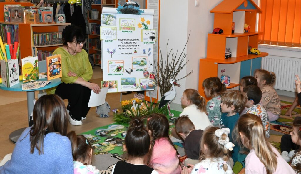 Bibliotekarka Irena Kozłowska siedzi przed dziećmi. Obok niej tablica z rysunkami, bazie w wazonie, stolik z książkami.
