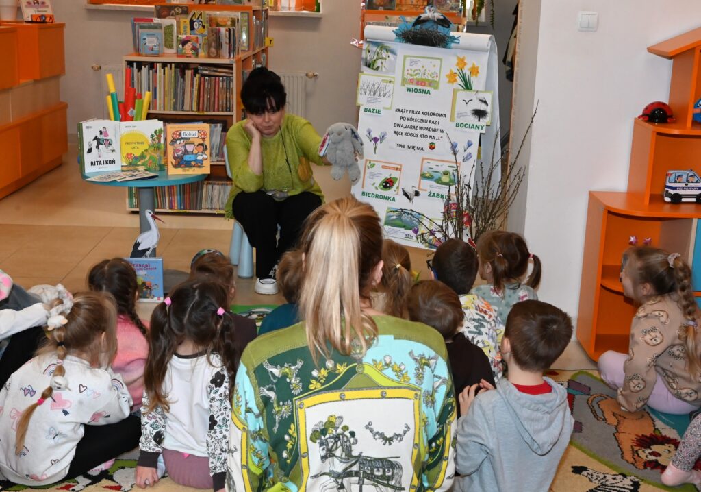 Bibliotekarka Irena Kozłowska siedzi przed dziećmi. trzyma w ręce pluszowego zająca. Obok niej tablica z rysunkami, bazie w wazonie, stolik z książkami.