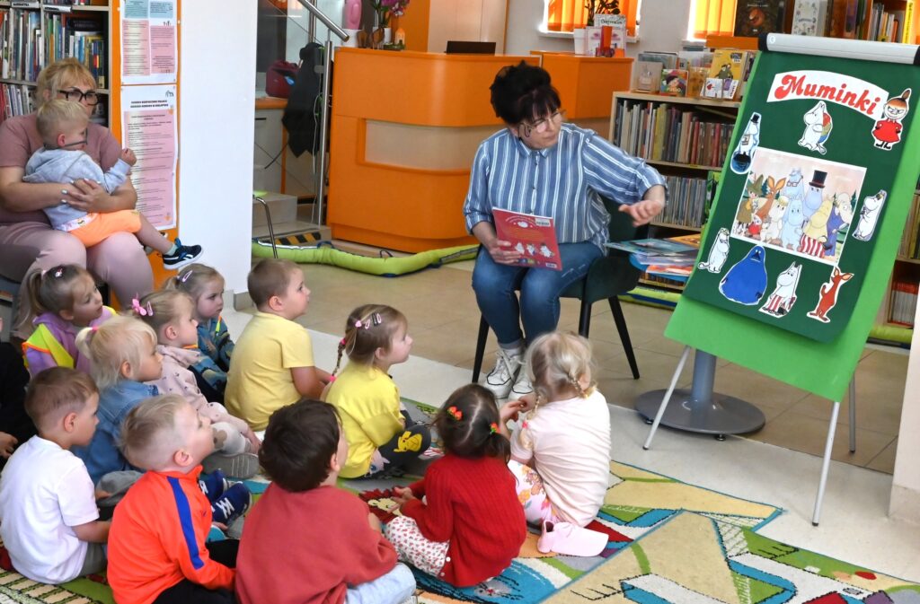 Bibliotekarka Irena Kozłowska trzyma w rękach książeczkę.. Obok niej na tablicy są postacie z "Muminków". 