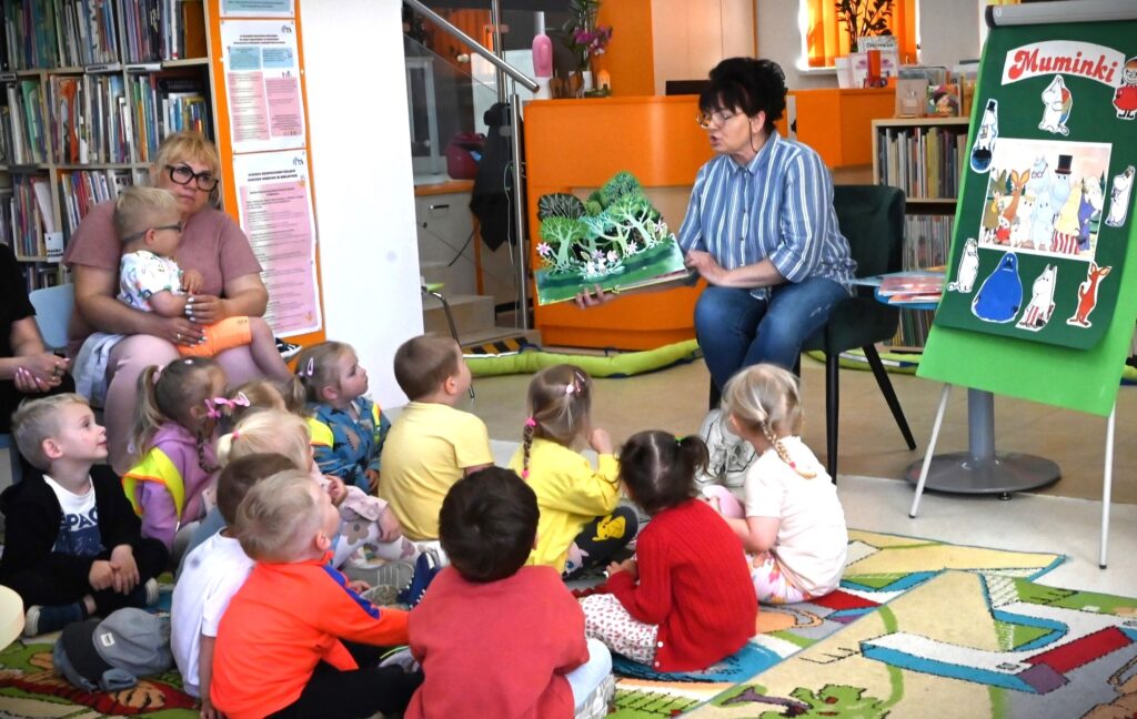 Bibliotekarka Irena Kozłowska pokazuje dzieciom rozkładaną książeczkę z drzewami. Obok niej na tablicy są postacie z "Muminków".