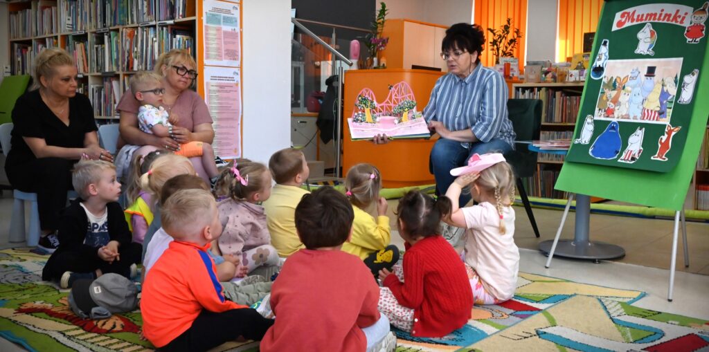 Bibliotekarka Irena Kozłowska pokazuje dzieciom rozkładaną książeczkę z mostem. Obok niej na tablicy są postacie z "Muminków".