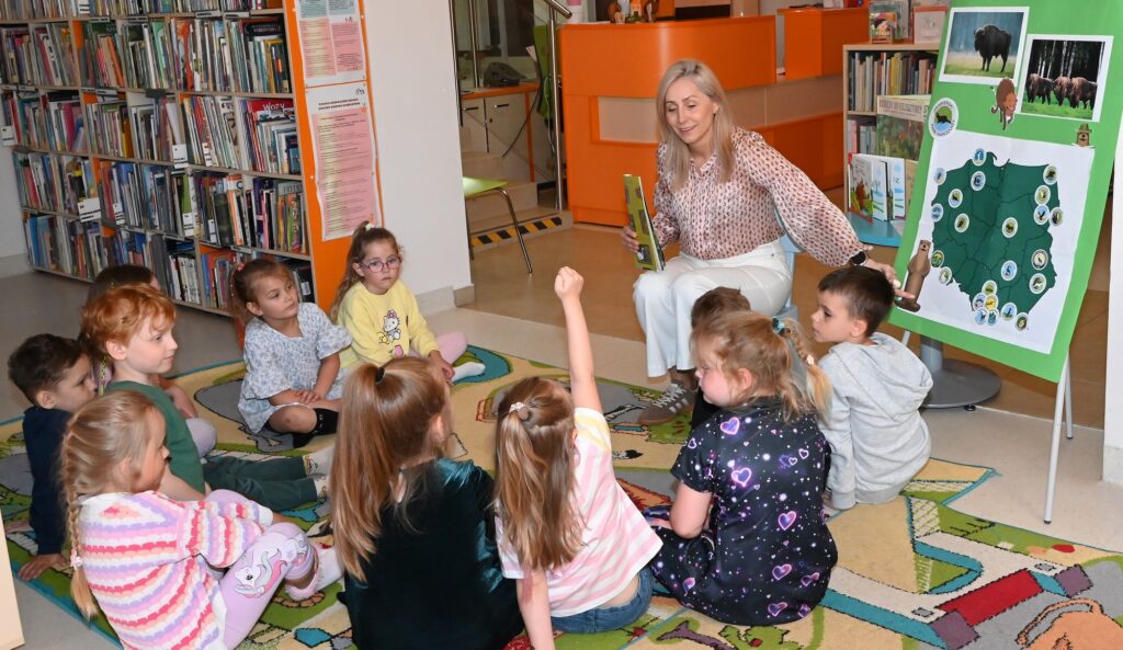 Przed bibliotekarką Izabelą Szczyż siedzą w kole dzieci, zaś obok niej stoi tablica, do której przyczepione są zdjęcia żubrów i wydry oraz mapa Polski, na której zaznaczone są parki narodowe.  Izabela Szczyż wskazuje na tablicy zdjęcie wydry. 