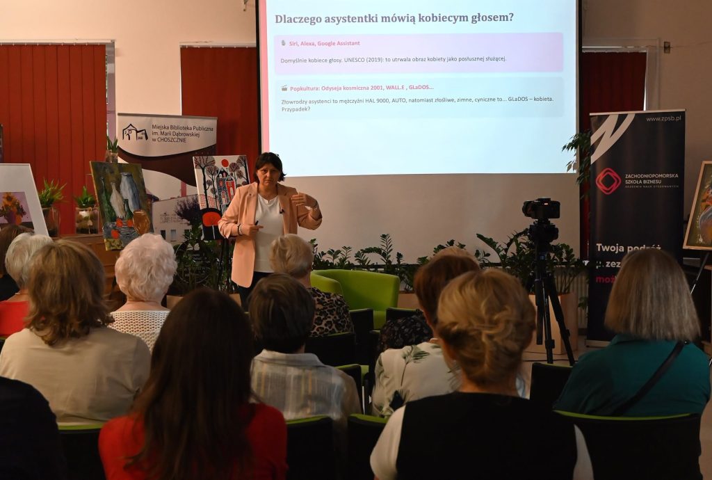 Dorota Dżega-Pietruszkiewicz przed publicznością. Z nią ekran z wyświetlonym slajdem "Dlaczego asystentki mówią kobiecym głosem?".