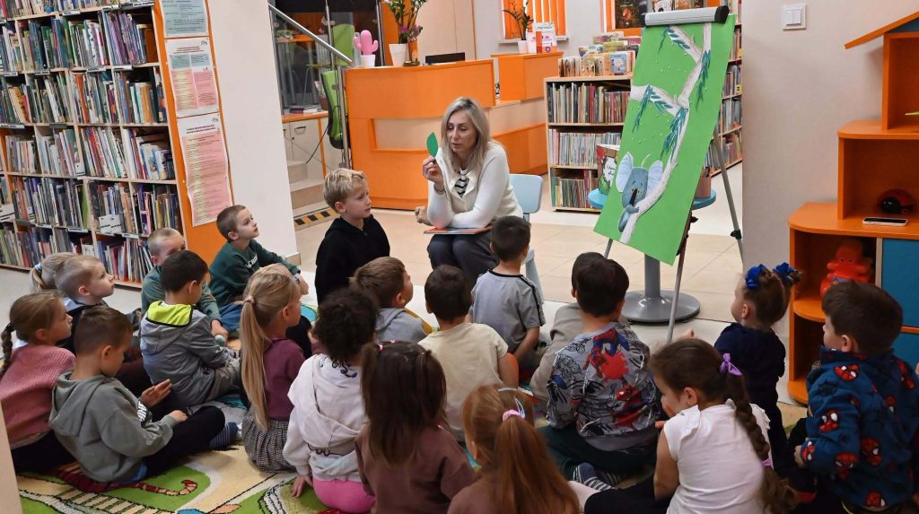 Bibliotekarka Izabela Szczyż trzyma w ręku wycięty z papieru liść i pokazuje go siedzącym na dywanie dzieciom. Obok na tablicy jest wyklejone drzewo i koala.