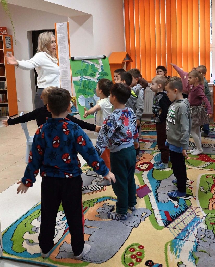 Bibliotekarka Izabela Szczyż i dzieci stoją na dywanie. Reę maja wyciągnięte na boki.