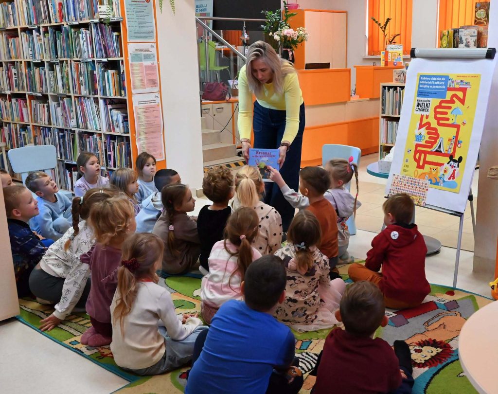 Bibliotekarka Izabela Szczyż pokazuje dzieciom siedzącym na dywanie książkę Część dzieci dotyka książki. obok niej stoi plakat "Mała książka, wielki człowiek".
