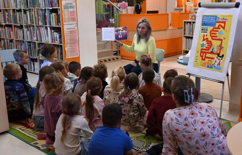 Bibliotekarka Izabela Szczyż pokazuje dzieciom siedzącym na dywanie otwartą książkę.