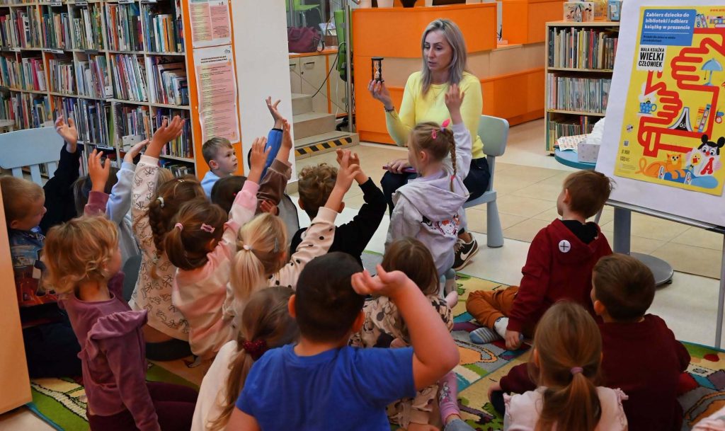 Bibliotekarka Izabela Szczyż pokazuje dzieciom siedzącym na dywanie klepsydrę.