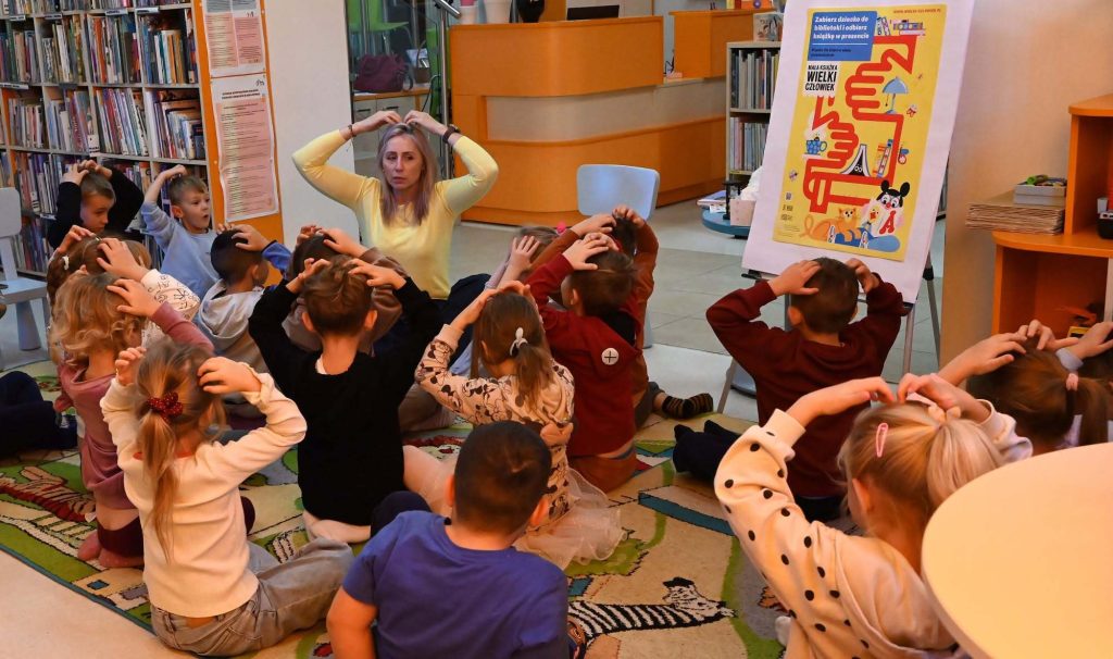 Bibliotekarka Izabela Szczyż oraz dzieci siedzące na dywanie dotykaj rękami swoich głów.