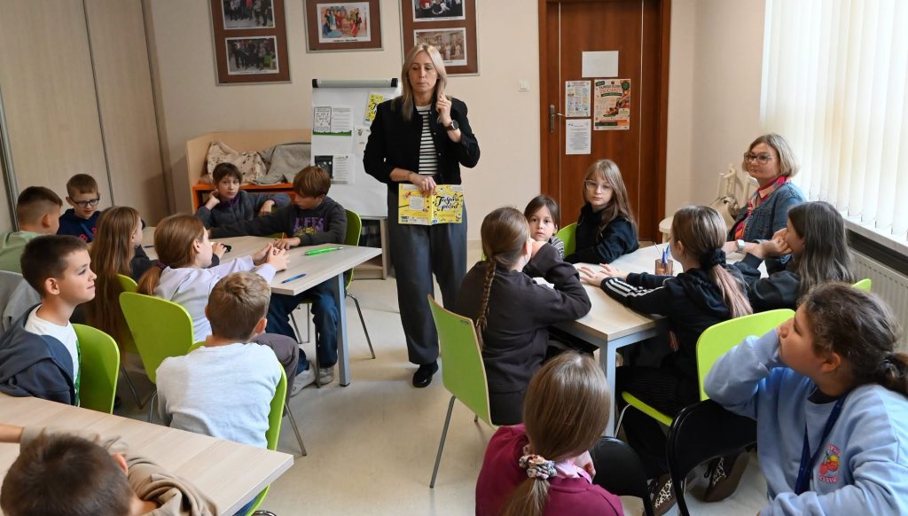 Bibliotekarka Izabela Szczyż stoi i trzyma w ręku obrazek. Obok stoi nauczycielka Za nią stoi flipchart Wokół przy stolikach siedzą uczennice i uczniowie, którzy zgłaszają się do odpowiedzi.