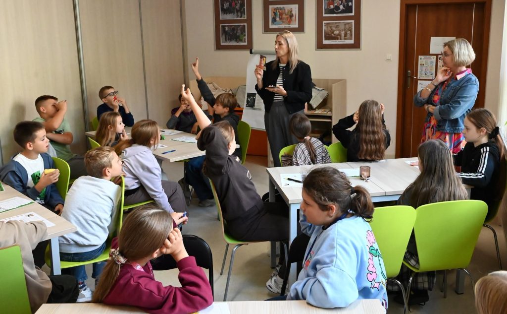Bibliotekarka Izabela Szczyż stoi i trzyma w ręku książkę. Za nią stoi flipchart Wokół przy stolikach siedzą uczennice, uczniowie i nauczycielka