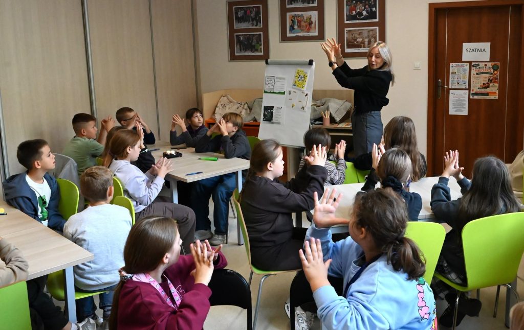 Bibliotekarka Izabela Szczyż stoi i trzyma w górze ręce. Obok stoi flipchart Wokół przy stolikach siedzą uczennice, uczniowie i nauczycielka, również podnoszą ręce.