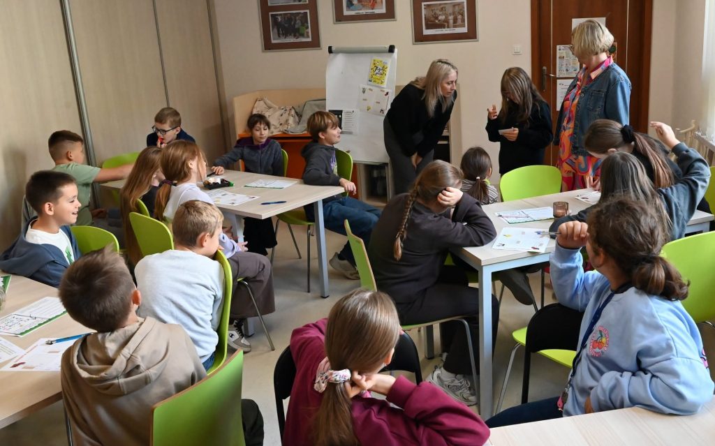 Bibliotekarka Izabela Szczyż pochyla się do uczennicy, która trzyma w ręku obrazek. Obok stoi nauczycielka.  Za nią stoi flipchart Wokół przy stolikach siedzą uczennice, uczniowie.