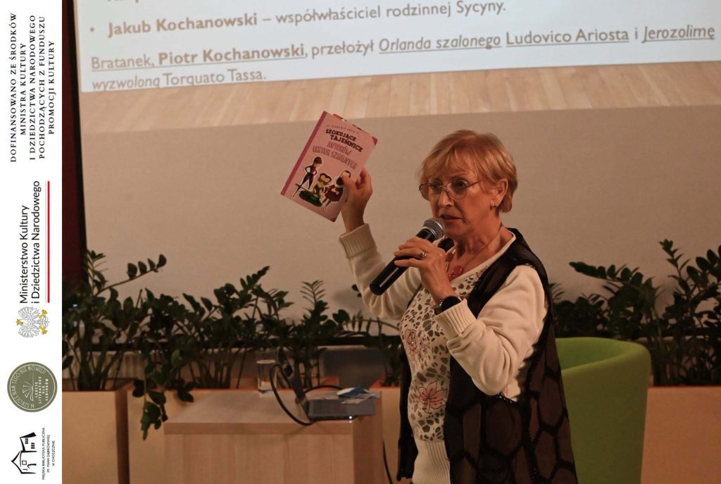 Cecylia Judek z książką w jednej ręce i mikrofonem w drugiej.