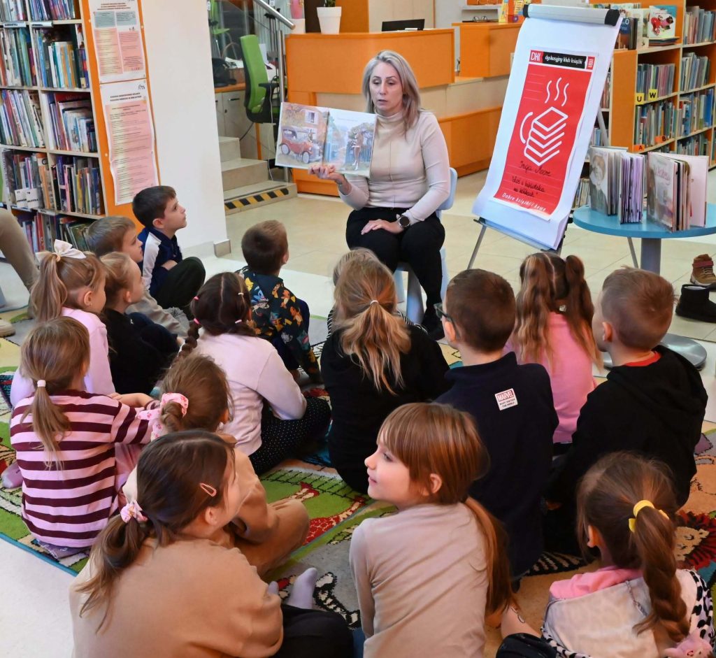 Bibliotekarka Izabela Szcyż pokazuje siedzącym przed nią na dywanie dzieciom książką. Obok stoi flipchart z plakatem DKKu.