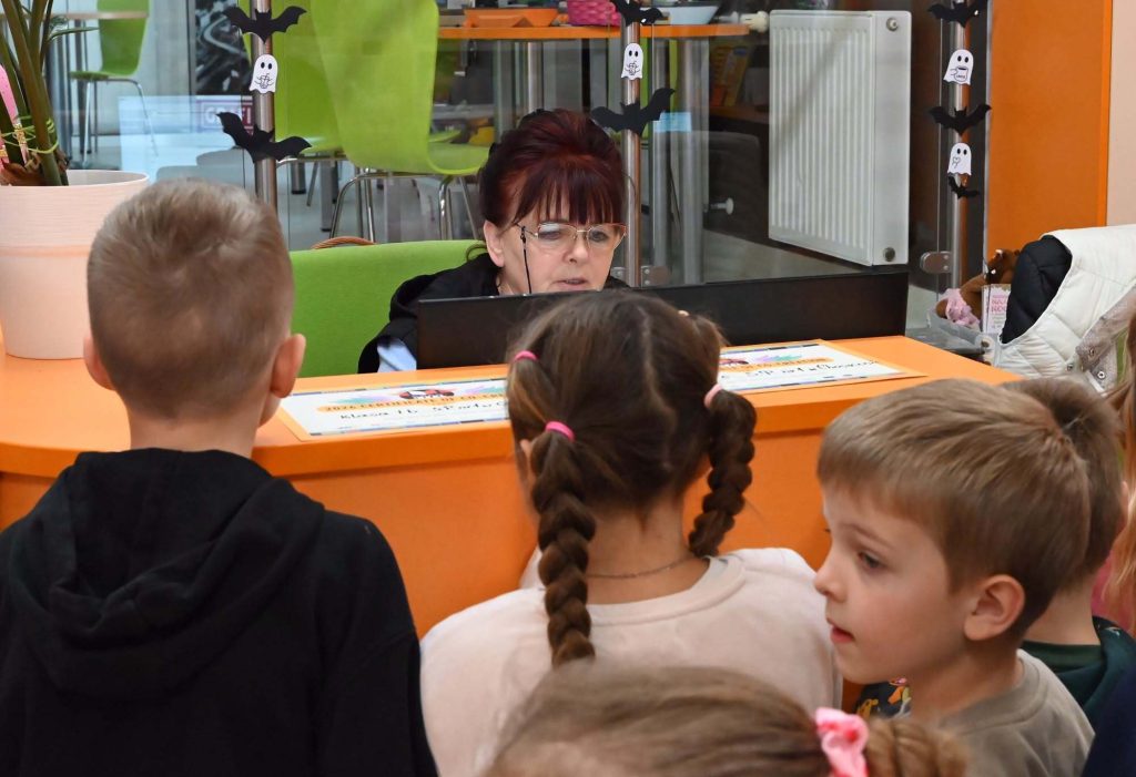 Za biurkiem siedzi bibliotekarka Irena Kozłowska i coś tłumaczy dzieciom.