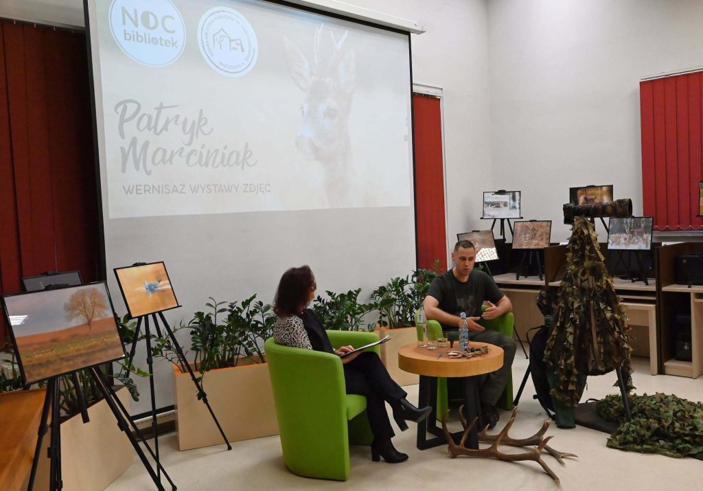 Od lewej siedzą Wioletta Witkowska i Patryk Marciniak. Za nimi slajd z tytułem spotkania. Przed stolikiem leży poroże jelenia. Obok stoi na statywie aparat fotograficzny, na nim wisi siatka maskująca.