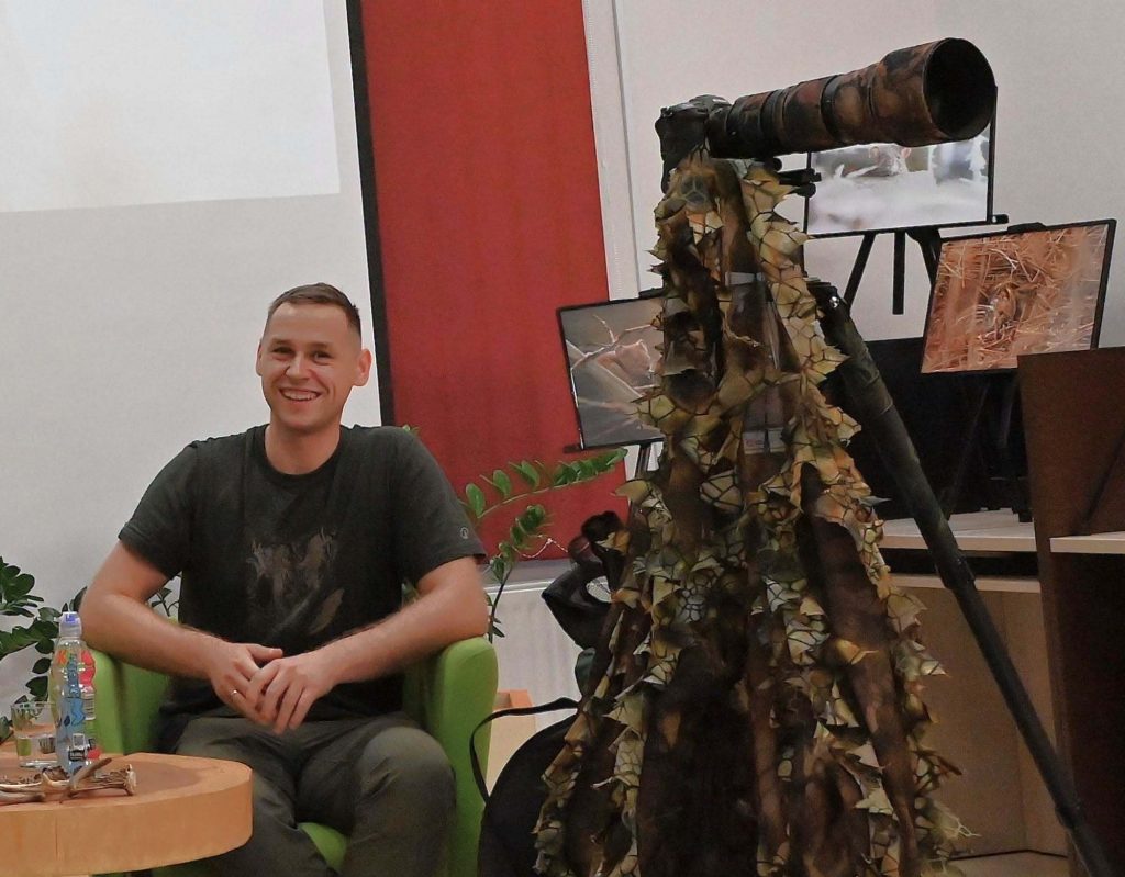 Patryk Marciniak. Obok stoi na statywie aparat fotograficzny, na nim wisi siatka maskująca.